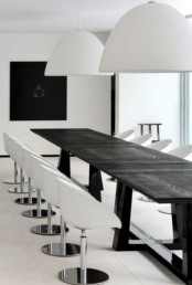 a minimalist black and white dining space with a long black table, white chairs, white pendant lamps and a black artwork on the wall