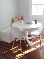 a very small and lovely white dining space with a corner bench, a round table, white chairs and printed pillows