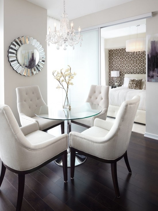 a glam dining space with a round glass table, white upholstered chairs, a crystal chandelier and a cool glam mirror is chic