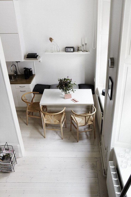 a small yet functional Scandinavian dining space with a bench with cushions, woven chairs, a built-in bench and a white table plus an open shelf over the space