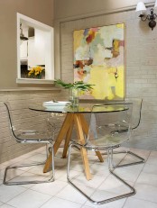 a small bold dining space with a glass table, acrylic chairs, a bold abstract artwork and some greenery and sconces is cool