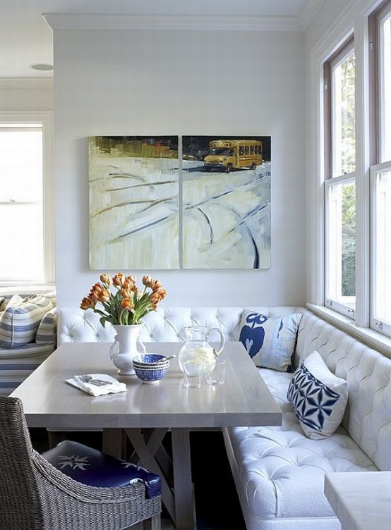 a small dining nook with a white upholstered corner bench, a grey table, a woven chair, some artworks and bold pillows