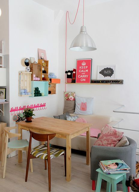 a colorful eclectic dining nook with a blush loveseat, a wooden table and mismatching chairs, crate shelves and an open shelf with artworks plus a pendant lamp on pink cord