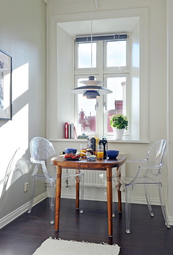 a cute little dining space wiht a stained table, ghost chairs, a pendant lamp and potted blooms and a view through the window