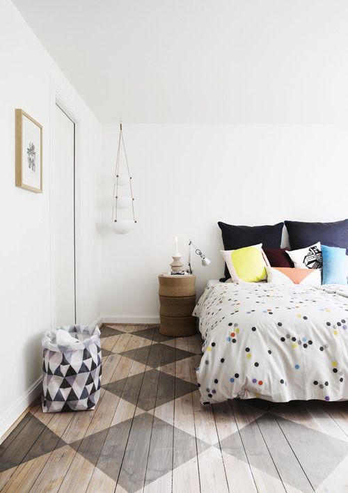 a neutral bedroom with a geo printed floor, a basket, a bed with polka dot and brigth bedding and a stacked box nightstand