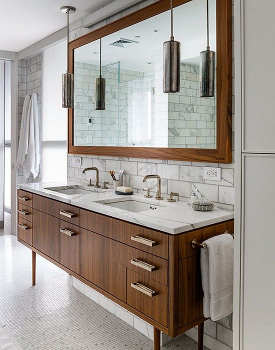 a refined mid-century modern bathroom clad with marble tiles, a wood frame mirror and a wooden vanity plus pendant lamps