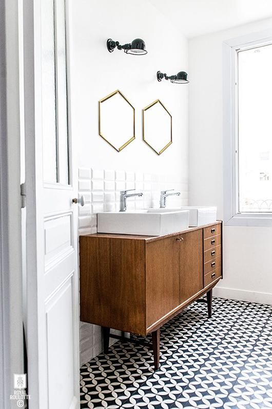 an elegant mid-century modern bathroom with black and white tiles on the floor, a mid-century modern vanity of wood, hex mirrors in gold frames