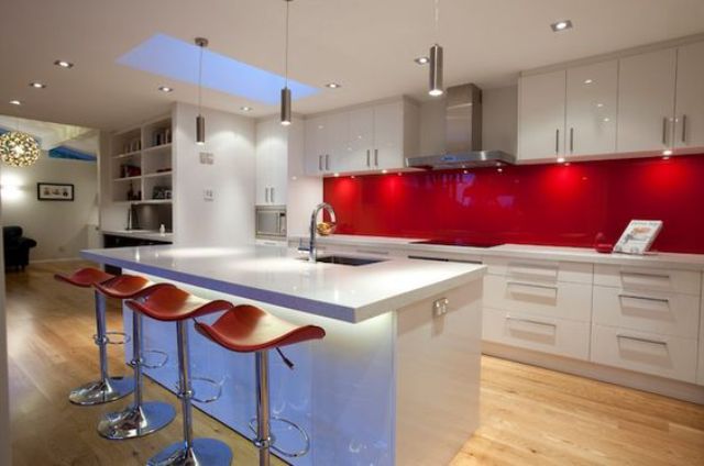 a modern white kitchen with sleek cabinets, a hot red glass backsplash, a lit up kitchen island and orange stools for a touch of color