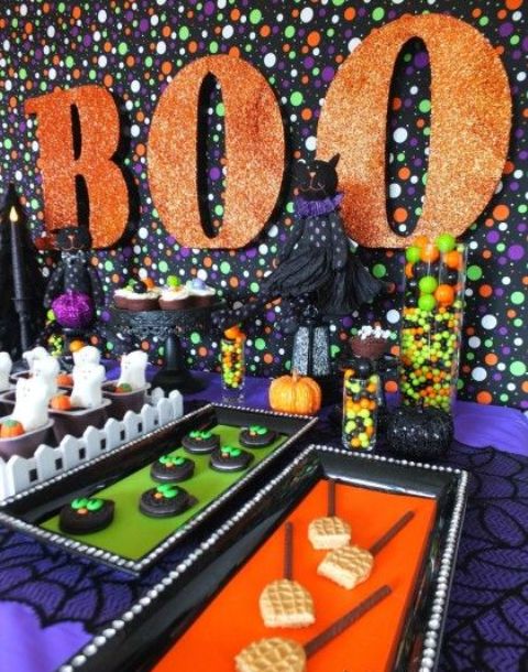 bright Halloween dessert table styling with a polka dot backdrop, orange glitter letters, bold sweets and candies on bold trays for a kids' party