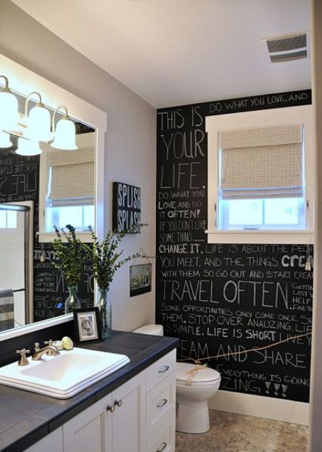 a creative black and white farmhouse bathroom with a chalkboard wall, a white vanity with a black countertop, a mirror with lamps, some art and greenery