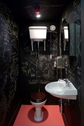 a catchy and bold powder room with black chalkboard walls, a wall-mounted sink, a toilet and a red floor plus some red touches