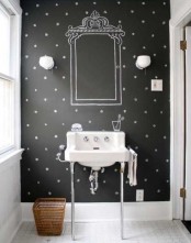 a black and white bathroom with a black chalkboard accent wall and a chalked mirror and decor, a sink on a stand, a basket and some sconces is a cool and modern space