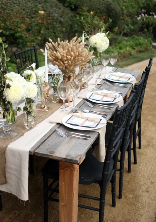 a rustic vintage Thanksgiving tablescape with a usual and burlap runner, a wheat and white bloom arrangements, white plates and neutral napkins, greenery and copper candleholders