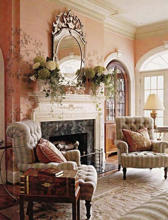 a sophisticated vintage living room with pink walls, a fireplace, a mirror in a chic frame, upholstered chairs, printed rugs and table lamps