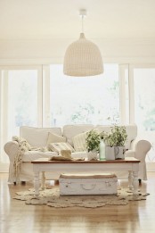 a neutral Provence-styled living room with a white sofa, a low coffee table and a suitcase, a woven pendant lamp and some potted greenery