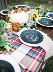 a Thanksgiving table setting with a plaid table runner, neutral linens, black plates, greenery, bold blooms and a pumpkin is a lovely idea to DIY
