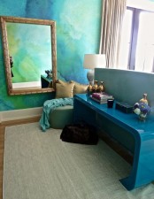 a bold space with a green and blue watercolor wall, a teal console table, a framed mirror and a green pouf with pillows
