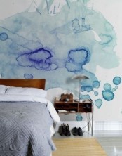 a bedroom with a light blue and bold blue watercolor accent wall, a stained bed with delicate blue bedding, a nightstand with a table lamp and a rug
