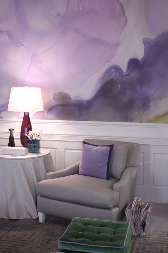 a chic space with a watercolor lilac accent wall, a grey chair with matching lilac pillows, a side table and a table lamp