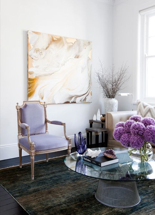 an airy refined living room with a lilac antique chair, a statement artwork, a dark rug and a glass coffee table with decor and flowers