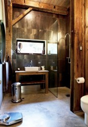 a rustic industrial bathroom clad with brown tiles and rough wood, with exposed wooden beams and a small window for a bit of light