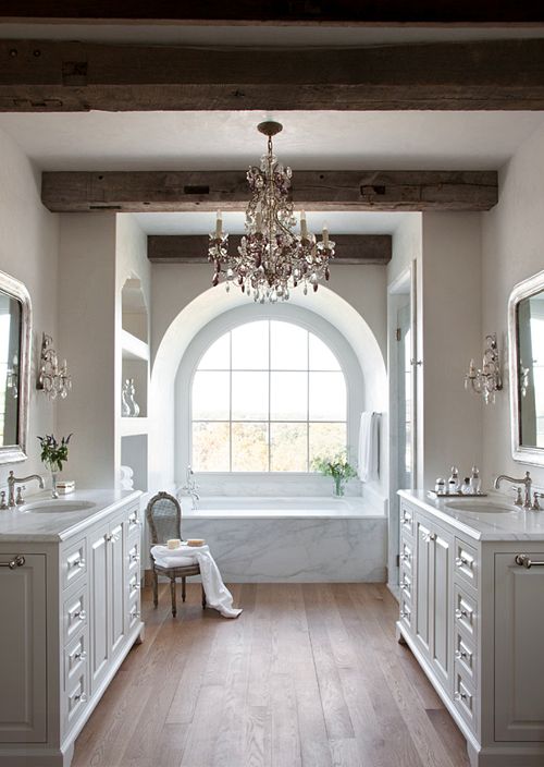 a refined bathroom with neutral walls, wooden beams on the ceiling, a refined crystal chandelier and white vanities