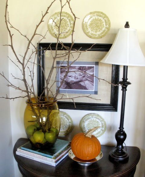 fall decor wiht a single pumpkin on a plate and some branches in a clear green vase plus apples inside it