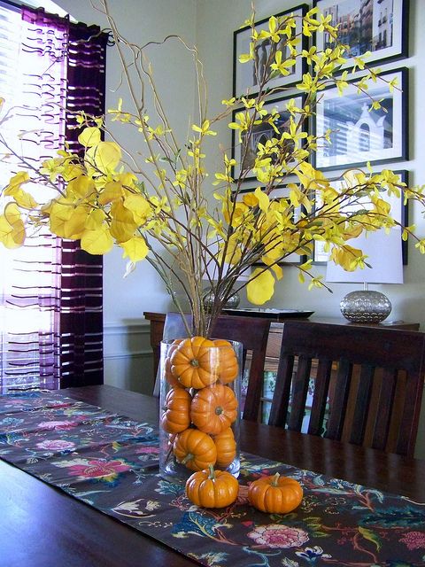 a stylish and simple fall centerpiece of a clear vase filled with orange pumpkins and branches with yellow leaves