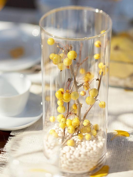 a fall centerpiece of a clear vase filled with white beads and with branches with yellow berries inside