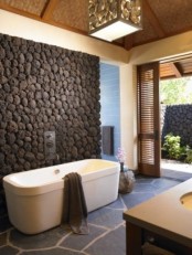 an outdoor-indoor contemporary bathroom with a stone floor and a decorative stone accent wall to make the bathtub stand out