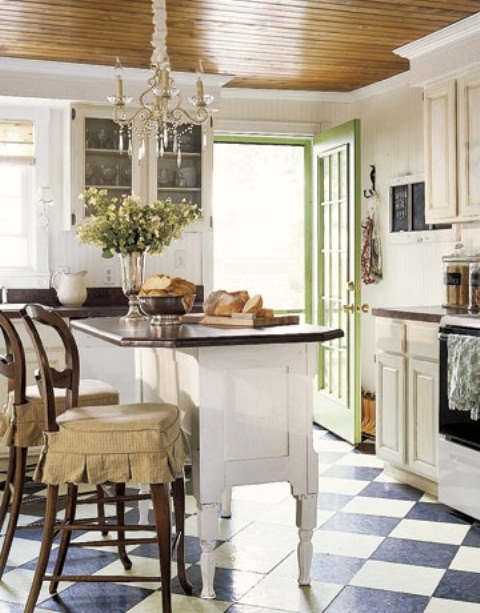 a white vintage kitchen with a checked floor, vintage white cabinetry, a white vintage kitchen island and vintage stained chairs