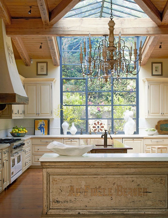 a large vintage kitchen in white and gold, with a large window and skylight, two vintage kitchen islands  and a large vintage chandelier over it