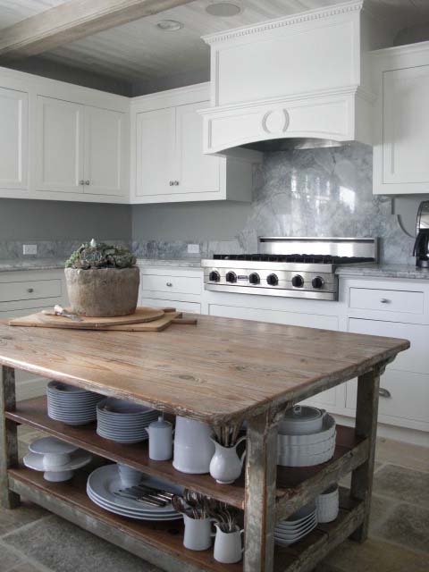 a vintage farmhouse white kitchen with a neutral stone backsplash and countertops, a large stained kitchen island that can double as a table