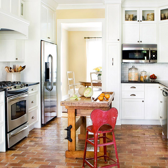 a white farmhouse kitchen with a brick floor, a small stained wood kitchen island, a red chair and black countertops for a touch of contrast here