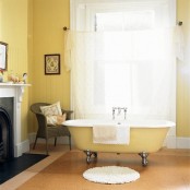 a cozy vintage sunny yellow bathroom with a non-working fireplace, a clawfoot bathtub, a curtain, artwork and a woven chair is very welcoming