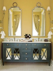 a chic bathroom with mustard walls, a navy double vanity, mirrors in eye-catchy frames and neutral textiles and sconces