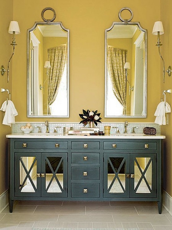 a chic bathroom with mustard walls, a navy double vanity, mirrors in eye-catchy frames and neutral textiles and sconces