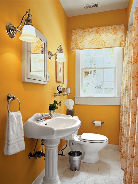 a small and coy country bathroom with bold yellow walls, a free-standing sink, wall sconces, artwork and some yellow floral curtains